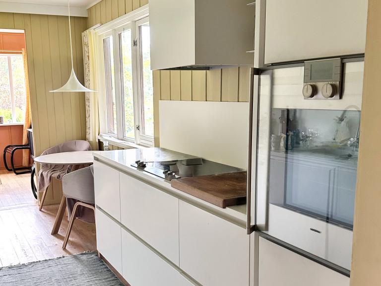 Kjøkken med peis. Benkeplater langs begge vegger. Gaggenau stekovn, dampovn og varmeskuff. Kokeplate, kjøleskap med frys. Miele oppvaskmaskin. I tillegg fryseboks i påbygg. Rundt spisebord med to stoler ved vindu følger med.